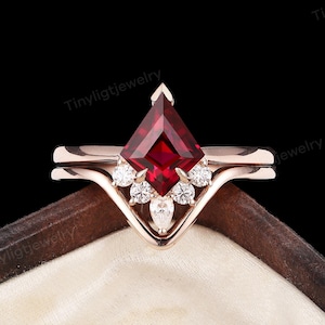 May include: A rose gold engagement ring set with a striking kite-cut, deep red gemstone as the focal point. The ring is embellished with small, round white gemstones and a marquise-cut stone. Presented in a wooden jewellery box.