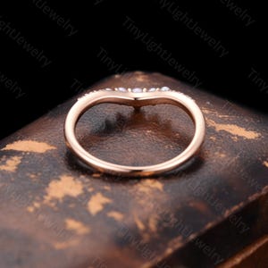 May include: A rose gold V-shaped ring featuring a curved band design. The top of the band is adorned with a series of small, round, white pearls set along the chevron-shaped curve, creating a delicate and symmetrical appearance on the metal band.