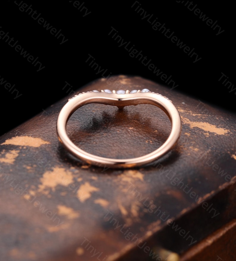 May include: A rose gold V-shaped ring featuring a curved band design. The top of the band is adorned with a series of small, round, white pearls set along the chevron-shaped curve, creating a delicate and symmetrical appearance on the metal band.