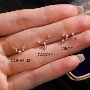 May include: Three rose gold stud earrings shaped like the Aquarius, Cancer, and Virgo constellations, each set with small clear stones. The text AQUARIUS, CANCER, and VIRGO is printed below each respective earring design.