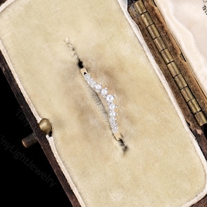 May include: A curved gold band ring featuring a row of alternating small white pearls and clear round-cut stones. The ring is set into a beige velvet-lined jewellery box with a hinged metal frame, displaying a delicate and symmetrical design.