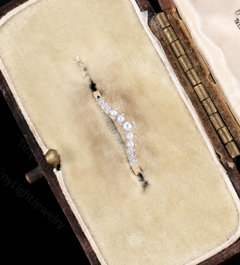 May include: A curved gold band ring featuring a row of alternating small white pearls and clear round-cut stones. The ring is set into a beige velvet-lined jewellery box with a hinged metal frame, displaying a delicate and symmetrical design.