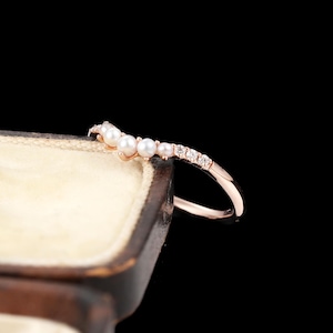 May include: A rose gold ring featuring a curved band set with a row of small white pearls and clear round-cut stones. The ring is positioned on the edge of a dark brown jewellery box with a cream-coloured interior against a black background.