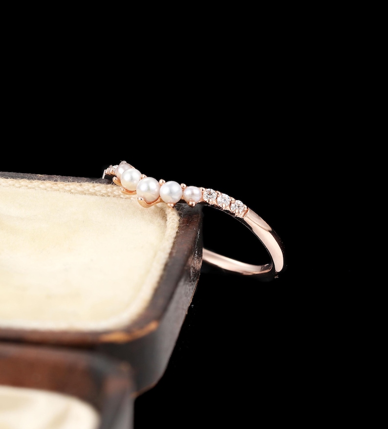 May include: A rose gold ring featuring a curved band set with a row of small white pearls and clear round-cut stones. The ring is positioned on the edge of a dark brown jewellery box with a cream-coloured interior against a black background.