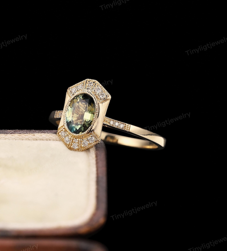 May include: A gold ring with an oval, green gemstone at the center. The ring has a geometric design with small, clear accent stones. The band is also adorned with small stones. The ring is displayed in a brown jewelry box.