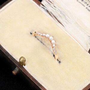 May include: A V-shaped ring crafted from rose-coloured metal, featuring a central arrangement of small white pearls flanked by clear, sparkling stones along the band. The ring is set into a cream-coloured velvet-lined jewellery box with a white tassel.
