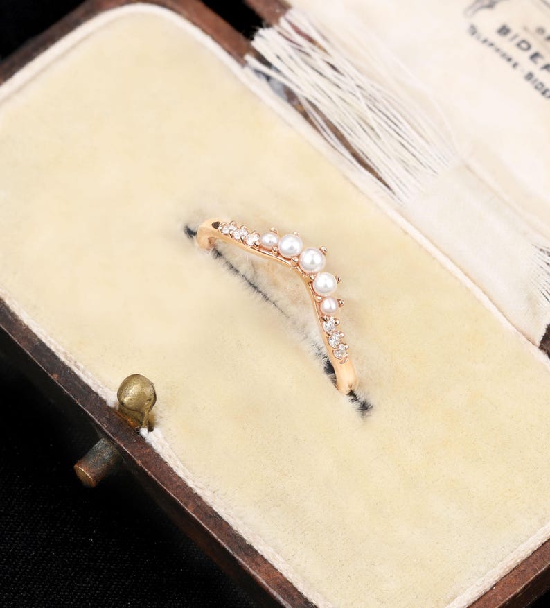 May include: A V-shaped ring crafted from rose-coloured metal, featuring a central arrangement of small white pearls flanked by clear, sparkling stones along the band. The ring is set into a cream-coloured velvet-lined jewellery box with a white tassel.
