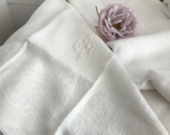 May include: White linen napkins with an embossed floral pattern and the initials "PB". A lavender rose rests on top of the folded napkins. The napkins are folded and stacked, suggesting a set.