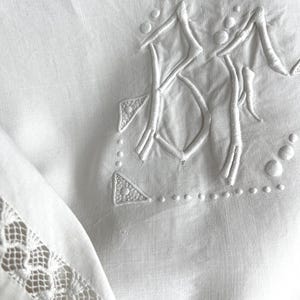 May include: Close-up of a white linen textile with embroidered details. The design includes a large, stylized monogram and decorative elements. A delicate lace trim adorns the edge of the fabric, adding a touch of elegance.