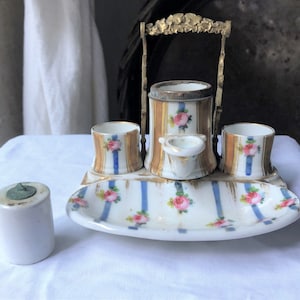 May include: Antique porcelain vanity set with a white base, gold accents, and pink rose floral designs. The set includes a tray, two small cups, a central container, and a small dish. A separate white cylindrical container with a green top is also present.