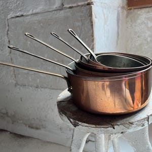 May include: A set of four nested copper saucepans with long, slender metal handles. The pans feature a polished, warm metallic exterior and a smooth, rounded silhouette, stacked neatly inside one another to display their varying sizes.