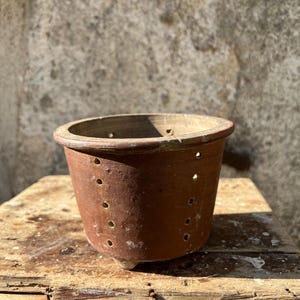 Op de afbeelding: Een rustieke terracotta plantenpot met drainagegaten. De cilindrische pot heeft een brede, afgeronde rand en staat op een verweerd houten oppervlak. De buitenkant van de pot vertoont tekenen van ouderdom en gebruik.