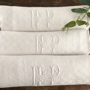 May include: Three white linen towels are stacked, each embroidered with the letters "MP" in a floral design. The towels are neatly rolled and arranged on a dark wooden surface, with a sprig of greenery in the background.