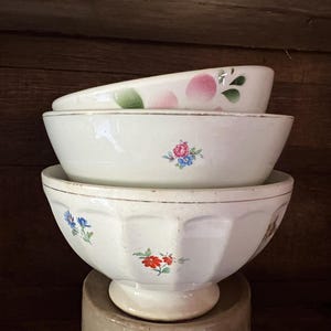 Op de afbeelding: Een stapel van drie witte keramische kommen met bloemmotieven. De bovenste kom heeft roze en groene accenten, de middelste kom heeft een klein bloemenboeket en de onderste kom heeft blauwe en rode bloemen.