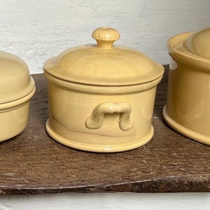 May include: Three matching yellow ceramic lidded containers with a matte finish. The containers are round with a small knob on top of the lid. The middle container has handles on the sides.