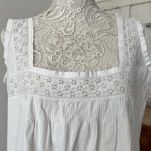 May include: A white sleeveless garment featuring a square neckline with intricate eyelet lace trim and floral embroidery. The bodice includes vertical pleats below the lace panel, and the fabric has a smooth texture. A matching smaller garment is visible.