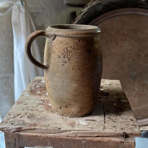 May include: A rustic, brown ceramic jug with a handle, set on a weathered wooden surface. The jug has a cylindrical body and a slightly flared rim. The background includes a woven tray and a white curtain.