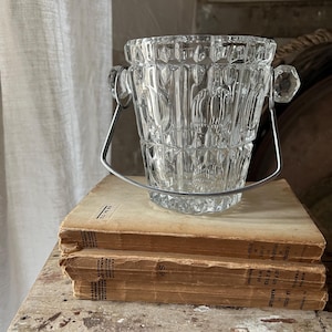 May include: A clear, faceted glass ice bucket with a silver handle and crystal accents. The bucket is sitting on top of two vintage books with aged, yellowed pages. The overall theme is vintage barware.