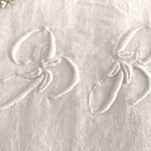 May include: Close-up of two white embroidered floral designs on a white textured fabric. The designs are detailed with curved lines and petal shapes, creating a delicate and elegant appearance. The overall theme is home decor.