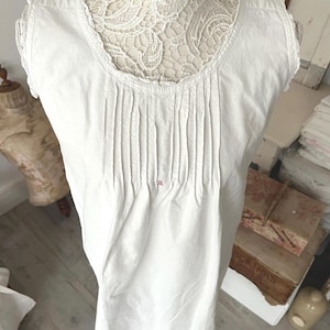May include: White sleeveless nightdress with a V-neck and vertical pleats. The neckline and shoulders are trimmed with lace. A small red embroidered detail is visible near the centre of the garment.