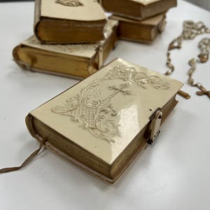 May include: Small ivory-coloured books with gold-gilded edges and embossed covers, featuring a cross design. A rosary with a cross and beads is also visible. The books are closed with a metal clasp and have a brown ribbon bookmark.