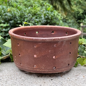 May include: A round, reddish-brown ceramic pot with evenly spaced holes around the sides. The pot sits on a concrete surface, with green foliage in the background. The pot has a rustic, aged appearance.