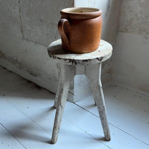 May include: A brown ceramic pot with a handle rests on a small, distressed white wooden stool. The stool has three legs and a round seat. The background shows a white wall and floor.