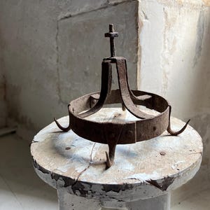 Peut inclure: Un trépied circulaire en fer rouillé doté de trois pieds incurvés et d'un montant central vertical surmonté d'un petit écrou. La surface métallique présente une oxydation importante et une texture rugueuse et patinée.