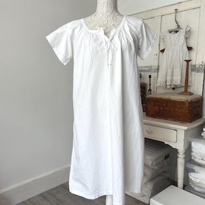 May include: A plain white cotton nightgown featuring short sleeves, a round neckline with a small front slit, and a loose, knee-length silhouette. The garment is displayed on a mannequin, showcasing a simple design with gathered fabric details.