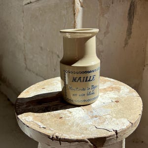 May include: A beige ceramic jar with the word "MAILLE" in blue script. The jar has a narrow neck and a wider base, sitting on a distressed, round, white stool. The text "Moutarde de Dijon au vin blanc" is also visible.