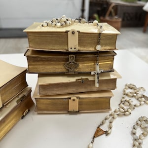 May include: A collection of antique books with aged, gold-edged pages and cream-coloured covers. A rosary with white beads and a cross hangs over the books. The books are secured with cream-coloured clasps.
