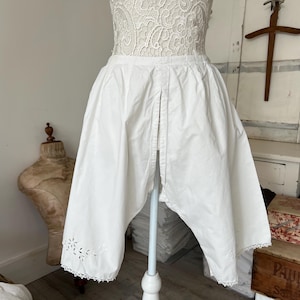 May include: White antique bloomers with a decorative floral cut-out design along the hem. The bloomers are made of a lightweight fabric and are displayed on a mannequin. The garment is part of a vintage clothing collection.