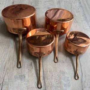 Può includere: Un set di tre pentole in rame lucidato con corpi arrotondati e lunghi manici robusti in ottone. Ogni pentola presenta una superficie metallica liscia e riflettente e una forma circolare distinta, disposte in ordine decrescente di dimensioni.