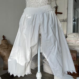 May include: White vintage bloomers with a wide leg and lace trim. The bloomers are displayed on a mannequin. The fabric appears to be cotton or a similar lightweight material. The garment has a high waist and a split front design.