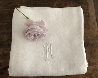 May include: White linen cloth with an embroidered letter "A" and a light purple rose. The cloth is folded and rests on a wooden surface. The rose is placed on the top left corner of the cloth.