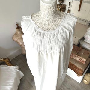 May include: A white sleeveless nightdress on a mannequin. The nightdress has a round neckline with lace trim and vertical pleats on the bodice. The mannequin's neck is decorated with detailed lace, and the garment is made of a soft, flowing fabric. The nightdress is a vintage style.