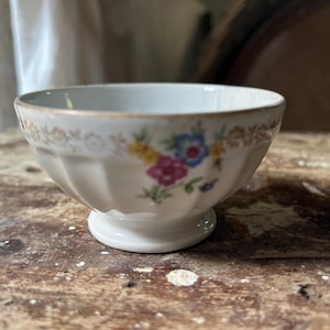 May include: A small, white, footed bowl with a floral design. The bowl has a scalloped edge and a gold trim. The floral design includes pink, blue, and yellow flowers. The bowl is sitting on a wooden surface.
