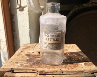 May include: A vintage clear glass bottle, labelled "BI-CARBONATE DE POTASSE". The bottle has a cylindrical shape with a wider base and a narrow neck. The label is aged and faded. It rests on a weathered wooden surface, suggesting age and use. The bottle is likely of French origin.