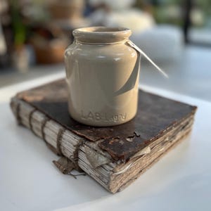 Peut inclure: Un pot en céramique beige avec l'inscription "LAB-Lagny" est posé sur un livre ancien et usé, avec une couverture en cuir marron et des pages vieilles. Une étiquette blanche est attachée au pot. Les objets sont sur une surface blanche.