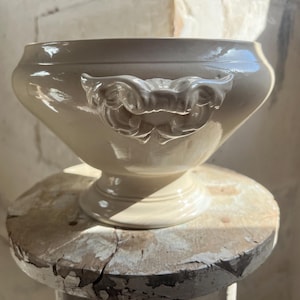 May include: A cream-coloured ceramic tureen featuring a wide, rounded bowl, a pedestal base, and two ornate, scrolled handles on opposite sides. The surface has a smooth, glossy finish that reflects light, highlighting the curved, sculptural design.