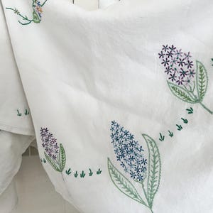 Può includere: Tessuto bianco con motivi floreali ricamati. Il ricamo presenta fiori di giacinto viola e blu con foglie e steli verdi. Sono ricamati anche piccoli dettagli verdi a forma di freccia.