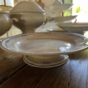May include: A vintage cream-coloured cake stand with a raised base and a wide, shallow plate. The plate has a blue and white decorative border. Other cream-coloured serving dishes are in the background, all on a wooden table.