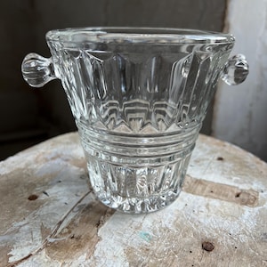 Könnte beinhalten: Ein klarer Eisbehälter aus Glas mit facettiertem Design und zwei kleinen, runden Griffen. Der Eimer hat eine breite Öffnung und eine leicht konische Form. Das Glas hat einen Vintage-Look und steht auf einer Holzoberfläche.