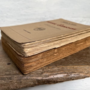 Può includere: Una pila di libri vintage con copertine e pagine colorate di marrone chiaro. Il libro in alto ha testo rosso e un emblema circolare. I libri poggiano su una superficie di legno stagionato, suggerendo età e utilizzo.