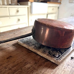 May include: A copper saucepan with a long, black handle rests on a decorative tile. The pan has a rounded body and a dark, aged appearance. The tile features a black and white floral pattern, set on a wooden table.