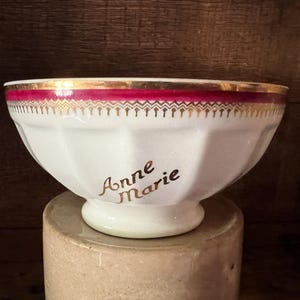 May include: White porcelain bowl with a gold rim and a red band with a gold pattern. The bowl has the name "Anne Marie" in gold script on the side. The bowl is sitting on a beige cylindrical base.