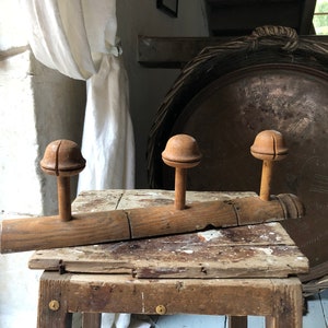 May include: A rustic wooden coat rack featuring three hat-shaped pegs. The rack is made of aged wood and rests on a weathered wooden table. A large, round metal tray and a white curtain are in the background.