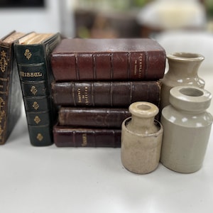 May include: A collection of antique books with leather covers in shades of brown and green, alongside small, beige ceramic jars. The books have gold lettering and decorative elements. The jars have a rustic, aged appearance.