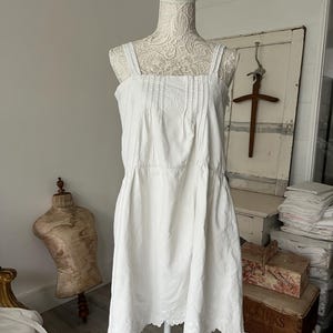 Könnte beinhalten: Weißes Baumwollkleid mit quadratischem Ausschnitt und dünnen Trägern, auf einer Schaufensterpuppe präsentiert. Das Kleid hat Falten am Oberteil und gestickte Details am wellenförmigen Saum. Das Kleid ist im Vintage-Stil.