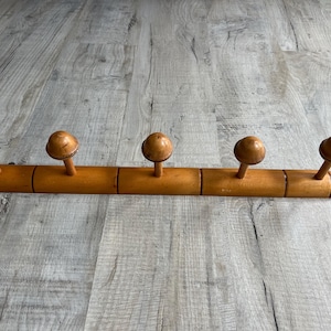 May include: A horizontal wooden wall rack featuring five evenly spaced pegs. Each peg is topped with a rounded, mushroom-shaped knob. The rack has a smooth, light brown finish with visible wood grain and decorative grooved detailing on the ends.